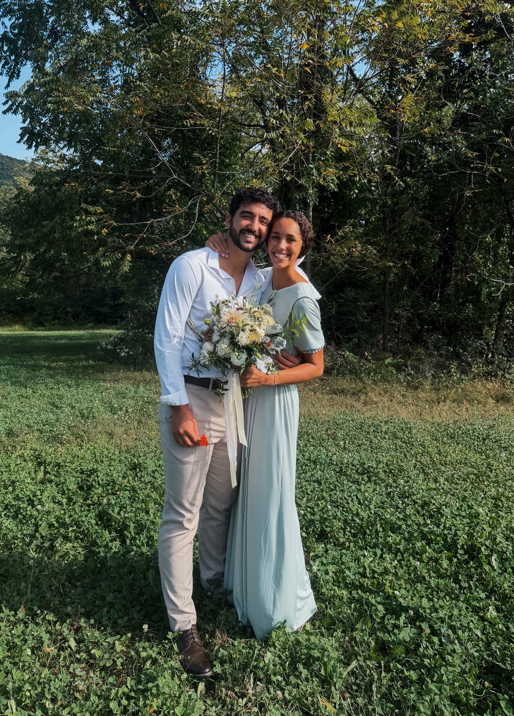 Kendall and Tirza Yoder smiling in a green field on their wedding day, Tirza holding a bouquet